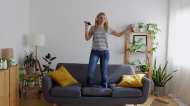 Evdeki oturma odasında kanepede dans ederken akıllı telefonuyla müzik dinleyen mutlu genç kadın. Boş zaman konsepti.
