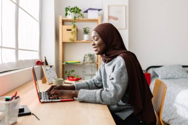 Evde dizüstü bilgisayar kullanan, başörtüsü takan mutlu siyah kız öğrenci. Genç Afrikalı bir kadın odasında öğreniyor ya da çalışıyor. Eğitim ve teknoloji kavramı.