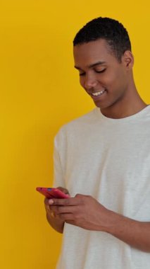 Dikey HD video. Sarı stüdyo arka planında cep telefonu kullanan gülümseyen genç z Afrikalı adamın yan görüntüsü. Teknoloji ve sosyal medya kavramı. 
