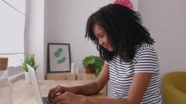 Young african american woman with curly hair working on her laptop computer while sitting on a desk at workplace. Happy millennial brazilian female studying online at home