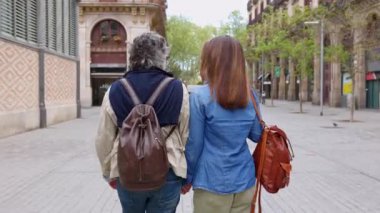 Şehir caddesinde yürüyen ve kameraya gülümseyen yaşlı turist çifti. Olgun çiftler Barselona 'yı ziyaret ederken el ele tutuşuyorlar. Emeklilik yaşam tarzı ve tatil konsepti.