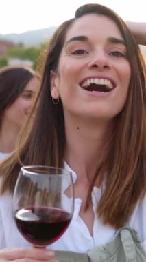 Dikey HD portre videosu. Mutlu güzel kadın kameraya gülümserken arka planda genç bir grup çok ırklı arkadaşla dans ediyor ve kırmızı şarap içiyor..