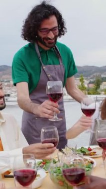 Yaz partisi kutlamalarında kırmızı şarap içen genç bir grup insanın dikey HD videosu. Çok ırklı arkadaşlar yemek masasında oturup terasta bbq yemeğinin tadını çıkarıyorlar..