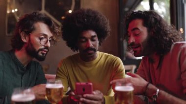 Bardaki masada oturan üç farklı yetişkin arkadaş cep telefonu kullanıyor. Teknoloji ve erkek arkadaşlığı kavramı.