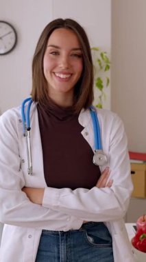 Vertical video portrait of young beautiful female nutritionist doctor standing over office workplace. Hospital worker in white coat looking at camera at clinical desk consultation. Healthcare concept