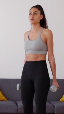Külotlu çorap ve üst giysili güzel genç bir kadının evde dambıllarıyla egzersiz yaptığı dikey HD videolar. Azim, spor ve sağlıklı yaşam tarzı kavramı. Ön görünüm.