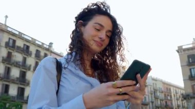 Akıllı telefonunu kullanarak Barcelona 'da şehir binalarının ve gün batımının başında duran güzel kıvırcık kızın portresi. Teknoloji yaşam tarzı ve kentsel konsept.