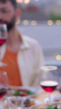 Dikey video. Bir grup genç, akşam çatı partisinde kırmızı şarap kadehleriyle tezahürat yapıyor. Çok ırklı arkadaşlar evde terasta bir araya gelip BQ yemeğini kutluyorlar.. 