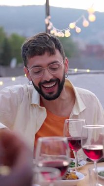 Dikey video. Genç yetişkin bir adam, arkadaşlarıyla dışarıda çatı katındaki masanın etrafında oturmuş, BBS yaz yemeğinin tadını çıkarıyor. Milenyum insanları kutlamayı ve eğlenmeyi seviyorlar..