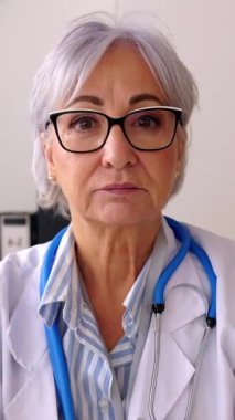 Üst düzey profesyonel sağlık görevlisinin kameraya baktığı dikey HD video. Tıbbi danışma masasında oturan olgun bir kadın doktorun kafa fotoğrafı. İlaç ve sağlık hizmetleri kavramı. 