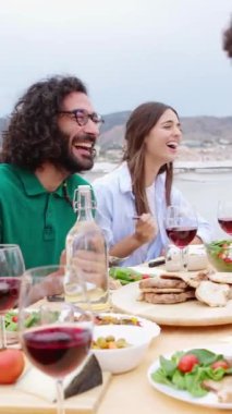 Dikey video. Genç ve mutlu arkadaşlar yaz partisi kutlamalarında birlikte yemek yiyorlar. Çok ırklı erkekler ve kadınlar barbekü yapmaktan ve terasta kırmızı şarap içmekten zevk alıyorlar..