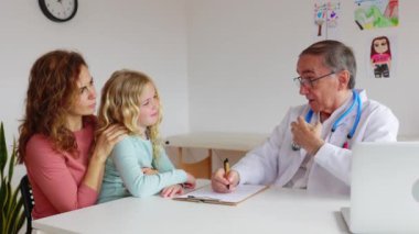 Doktor muayenehanesinde anne ve kız çocuğuyla konuşan bir çocuk doktoru. Tıbbi konsültasyondaki hastaya reçete yazan kıdemli sağlık görevlisi. Aile hekimi konsepti.