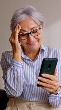 Evde dinlenirken cep telefonu kullanan mutlu yaşlı bir kadının dikey HD videosu. Gülümseyen, 60 'lı yaşlarda beyaz saçlı, akıllı telefon ekranına bakan bir kadın. Teknoloji ve yaşlılar konsepti.
