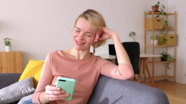 Orta yaşlı bir kadın cep telefonu kullanarak koltukta dinleniyor. Mutlu Kuzey Avrupalı kadın cep telefonu uygulamasından sosyal medya içeriğini izlerken eğleniyor. Yüksek kalite 4k görüntü