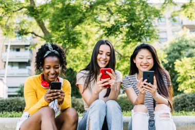 Yazın şehir parkında oturan üç tane çok ırklı kadın cep telefonu kullanıyor. Teknoloji ve sosyal medya kavramı.