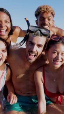 Dikey video. Suda şişme minderleri olan çok ırklı arkadaşların komik grup portresi sahilde yaz tatilinin tadını çıkarıyor. Tatil yaşam tarzı ve arkadaşlık kavramı.
