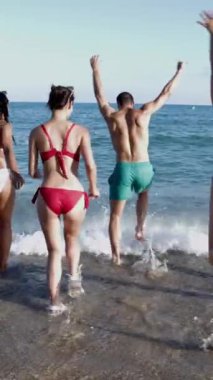 Dikey video. Yaz tatilinin tadını çıkaran çeşitli genç arkadaş gruplarının yaz mevsiminde denize doğru koşuşlarının arka görüntüsü. Turist sahilde tatilde eğleniyor..