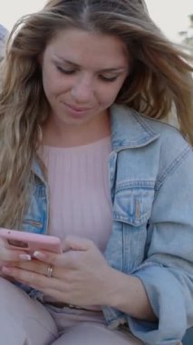 Dışarıda arkadaşlarıyla oturan akıllı telefon kullanan güzel bir kadının dikey HD videosu. Teknoloji ve sosyal medya kavramı.