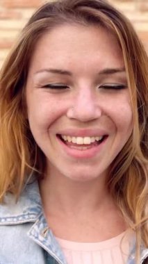 Dikey video. Duvarın üzerinden kameraya gülümseyen güzel sarışın genç bir kadının açık hava portresi. Mutluluk ve mutlu insanlar konsepti.