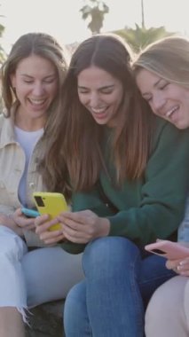 Dikey video. Dışarıda cep telefonu kullanan genç bayan arkadaşlar. Milenyum kadınları akıllı telefon uygulamasında sosyal medya içeriğini izlerken eğleniyorlar. Teknoloji ve gençlik toplumu kavramı.
