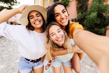 Yaz tatilinde cep telefonuyla fotoğraf çektirerek eğlenen üç mutlu genç kadından oluşan çeşitli bir grup. Seyahat ve tatil yaşam tarzı kavramı.