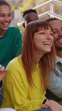 Bir grup genç arkadaş dışarıda birlikte eğleniyor. Millennial çok ırklı insanlar şehrin merdivenlerinde sosyal bir toplantı yaparken gülüyorlar. Arkadaşlık kavramı. Dikey HD video.