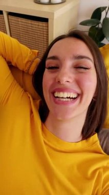 Evdeki kanepede yatarken cep telefonuyla video görüşmesi yapan mutlu genç bir kadının dikey HD videosu. İletişim konsepti.