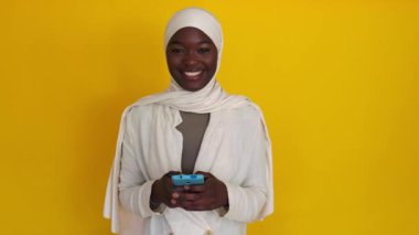 Cep telefonlu Afrikalı Müslüman kadın sarı arka planda kameraya gülümsüyor. Teknoloji ve sosyal medya kavramı.