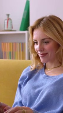 Evdeki kanepede otururken dizüstü bilgisayar kullanan mutlu sarışın kadının dikey HD videosu. Milenyum kız öğrencileri evde internetten öğreniyor..