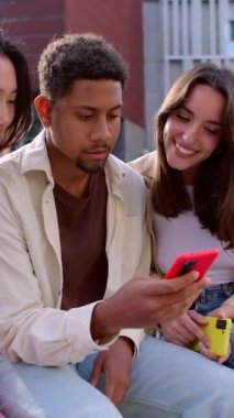 Açık havada cep telefonu kullanarak eğlenen genç nesil çoklu etnik grupların dikey HD videosu. Gülümseyen öğrenci topluluğu ellerinde akıllı cep telefonlarıyla şehirde birlikte oturuyorlar.