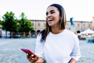 Şehir caddesinde cep telefonu kullanan mutlu, çok ırklı genç bir kadın. Neşeli Afrikalı Amerikalı kız dışarıda akıllı telefonu tutarken gülüyor. Teknoloji yaşam tarzı konsepti.
