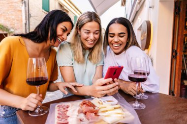 Barda aperatiflerin tadını çıkarırken cep telefonu kullanan üç genç kadın. Milenyum çeşitliliğindeki İtalyan kızları kırmızı şarap içerken akıllı telefondan sohbet ediyorlar.