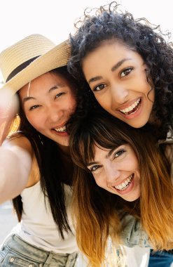 Şehir caddesinde eğlenen üç çok ırklı genç kadının dikey fotoğrafı. Milenyum arkadaş grubu yaz tatilinin tadını çıkarıyor açık havada selfie çekiyorlar. Arkadaşlık ve gençlik kavramı.