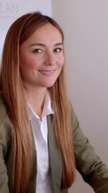 Vertical video. Cheerful portrait of young businesswoman looking at camera sitting in the office. Successful portrait of european professional female worker at workplace.