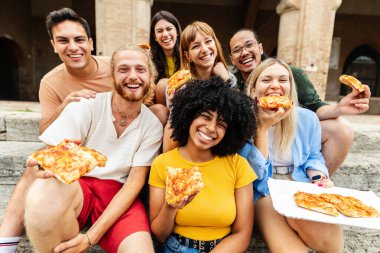 Dışarıda pizza yerken kameraya gülümseyen bir grup genç arkadaş. Şehir caddesinde dinlenirken İtalyan fast food 'unun tadını çıkaran çok ırklı mutlu insanlar. Seyahat ve yaz yaşam tarzı konsepti.