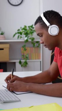 Genç siyahi adam dizüstü bilgisayarda çalışıyor ve not defterine not alıyor. Zombi çocuk internetten ders çalışıyor, Webinar izliyor, Webinar kurslarını dinliyor. Eğitim ve e-öğrenme konsepti. Dikey video.