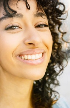 Dışarıdaki kameraya gülümseyen genç kadının dikey yakın portresi. Neşeli esmer, bin yıllık dişlek gülüşlü kadın. Şehir caddesinde kameraya bakıyor.