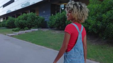Kentin caddesinde afro saçlı, kendinden emin, genç bir Latin Amerikalı kadın yürüyor. Yan görüş. 4K video.
