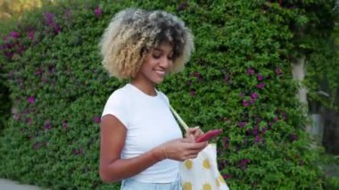 Genç Afro-Amerikan bir kadının şehir caddesinde cep telefonu kullanışının yan görüntüsü. Z Gen kızı yazın dışarıda akıllı telefon uygulamasından mesaj yolluyor. Teknoloji yaşam tarzı ve sosyal medya kavramı.