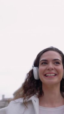 Şehrin sokaklarında dans ederken internetten müzik dinleyen neşeli genç bir kadının dikey HD videosu. Tasasız ve mutluluk konsepti.