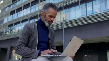 Bazı ofis binalarının yakınında dizüstü bilgisayarda çalışan kıdemli Afro-Amerikan iş adamı. Başarılı, olgun, profesyonel işçi şehir parkında oturan bilgisayardaki e-postaları kontrol ediyor.. 
