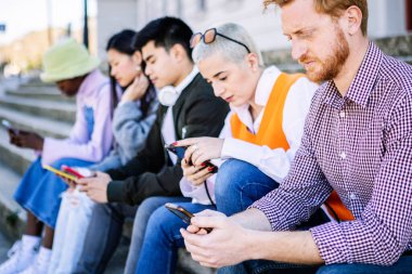 Dışarıda oturan akıllı telefonları kullanan çok ırklı genç arkadaşlar. Milenyum çeşitliliğindeki yetişkin insanlar cep telefonlarına bağımlılar ve sosyal medya içeriğini izliyorlar.