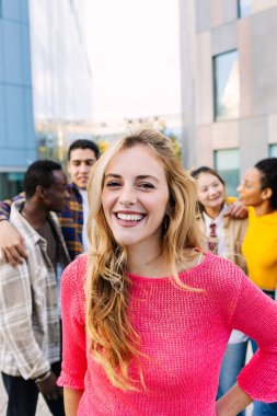 Güzel sarışın bir kadının dikey portresi bir grup arkadaş üzerinden kameraya gülümsüyor. Gençlik toplumu ve arkadaşlık kavramı.