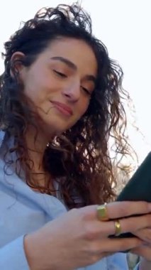 Güzel kıvırcık kızın dikey HD portresi Barcelona 'da şehir binalarının ve gün batımının başında duran akıllı telefonunu kullanıyor. Teknoloji yaşam tarzı ve kentsel konsept.