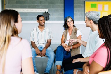 Young group of teenage students discussing in classroom sitting in circle with female teacher, giving an opinion on a debate topic. Education concept clipart