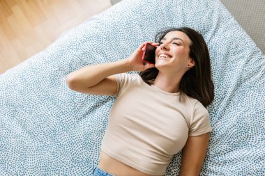 Yatakta uzanmış, cep telefonuyla konuşan mutlu genç bir kadın. Milenyum kadını cep telefonuyla arkadaşlarıyla sohbet ediyor..