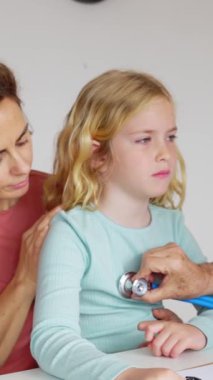 Pediyatrist kıdemli doktorun, muayene odasında küçük bir kızın kalp atışlarını ve nefesini dinlerken çekilmiş dikey HD videosu. Çocuk sağlığı konsepti.
