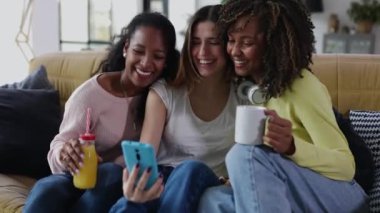 Evde birlikte cep telefonu kullanan çok ırklı genç bayan arkadaşlar. Mutlu kadın milenyum arkadaşları birlikte eğleniyor ve eğleniyorlar. Arkadaşlık ve teknoloji kavramı.