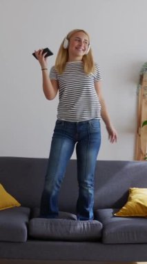 Evdeki oturma odasındaki kanepede dans ederken akıllı telefonlu müzik dinleyen mutlu genç kadının dikey HD videosu. Boş zaman konsepti.