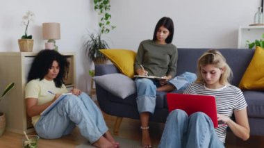 Üç genç kız öğrenci oturma odasında birlikte ders çalışıyor. Millennial bir grup kadın öğrenci aynı apartmanda üniversite atamaları üzerinde çalışıyor. Eğitim yaşam tarzı konsepti.